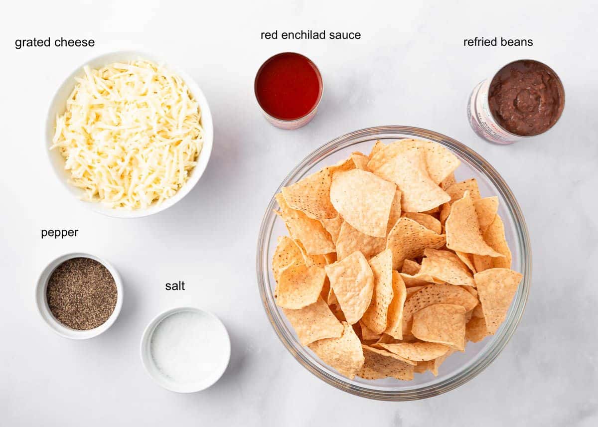 Ingredients for oven baked nachos including tortilla chips, shredded cheese, red enchilada sauce, refried beans, salt and pepper.
