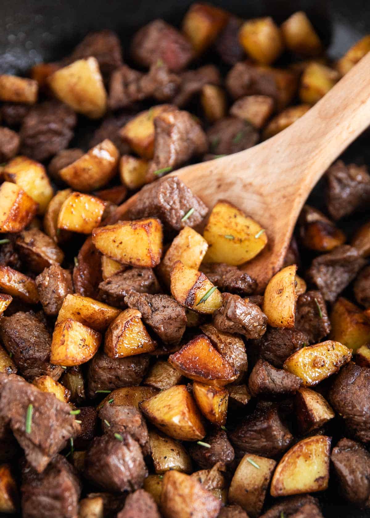 Steak and potatoes in a skillet with a wooden spoon.