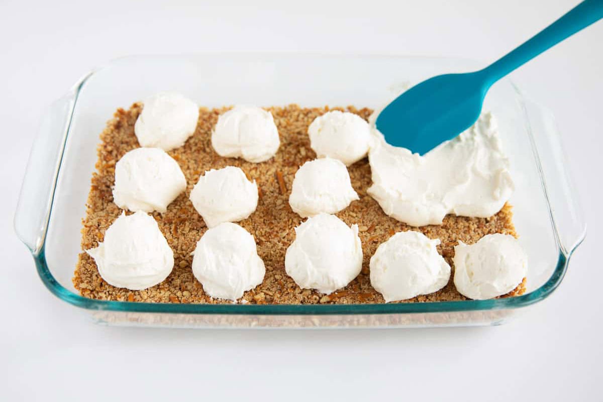 Spooning cream cheese filling over a baked pretzel crust in a 9x13 glass baking dish,