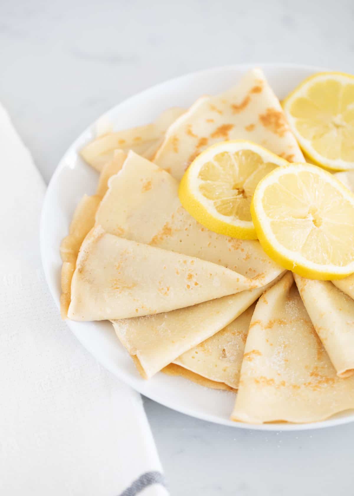 These deliciously simple lemon crepes are light and airy with a crispy outer edge and a zesty lemon flavor. They are perfect drizzled with lemon juice and powdered sugar or filled with whipped cream and lemon curd! Lemon crepes on white plate.