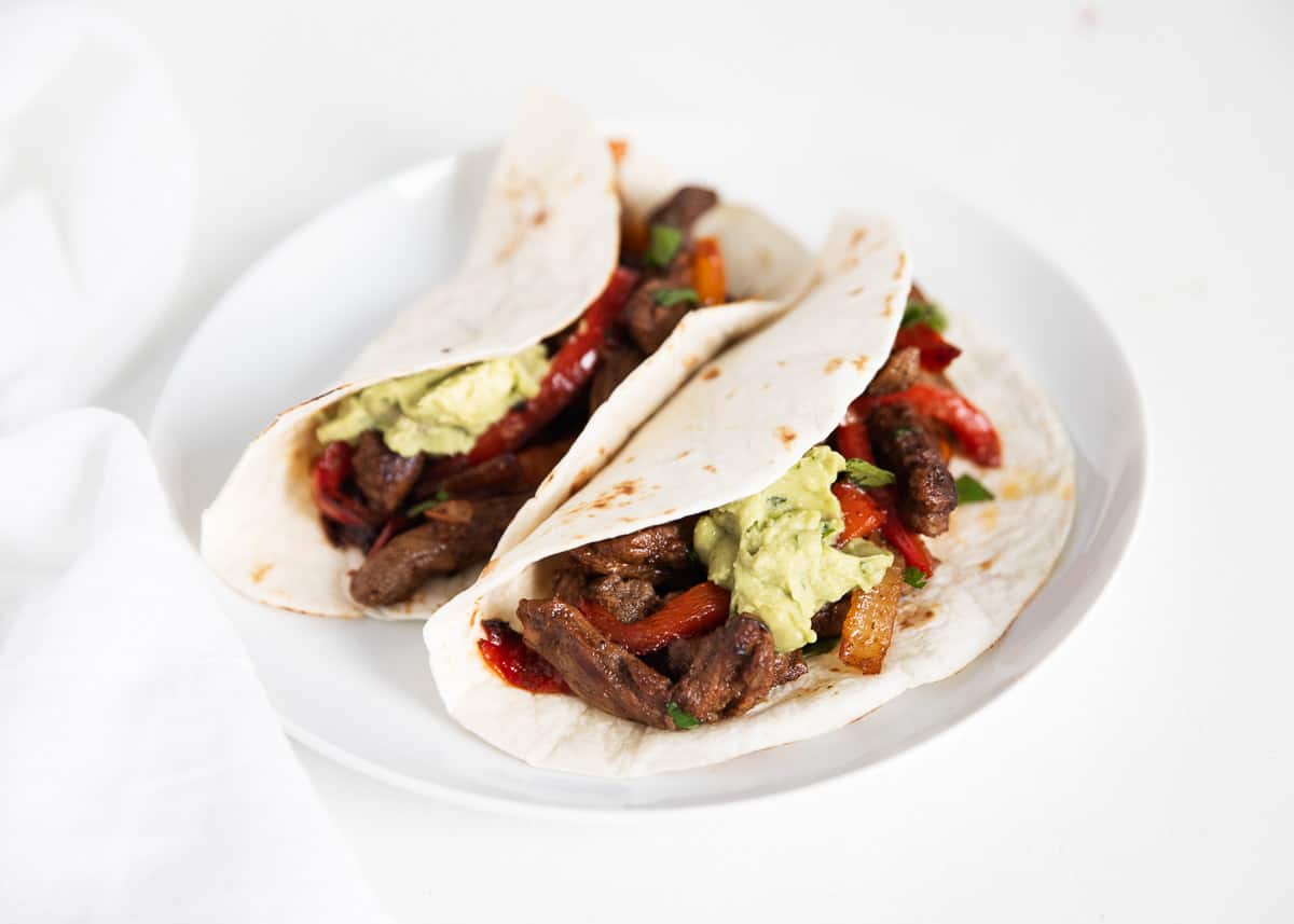 These mouthwatering steak fajitas are made with marinated strips of juicy steak, sautéed onions and peppers, and filled to the brim with all of your favorite zesty toppings! Steak fajitas on white plate.