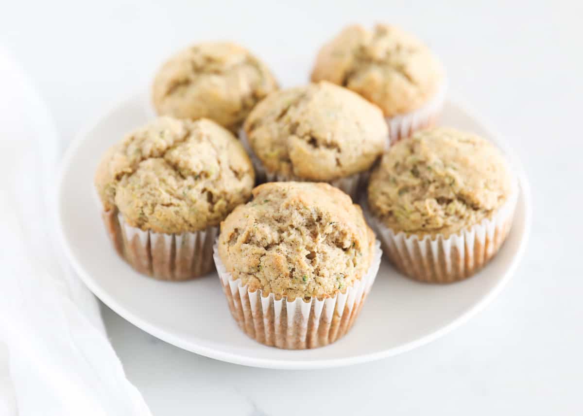 The best zucchini muffins are moist, fluffy, and loaded with the mild flavor of freshly grated garden zucchini! Flavored with a hint of cinnamon and vanilla, this classic recipe is easy to make in one bowl. Freshly baked zucchini muffins on a white plate.
