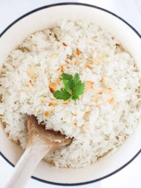 Coconut rice in a white pot with wooden serving spoon and coconut flakes with cilantro garnish.