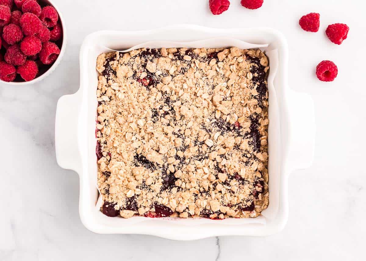 Chewy raspberry crumble bars made with a buttery oatmeal base, fresh raspberry jam and then finished off with a sprinkle more of the oatmeal crumb topping. You only need 7 ingredients and 15 minutes of prep time to make this easy summer dessert! Raspberry crumb bars in white pan.