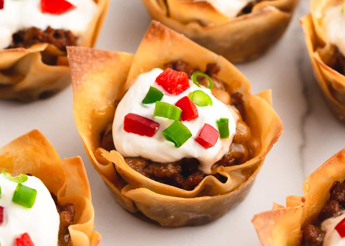 Layers of crispy wonton wrappers, seasoned taco meat with chili beans, melted cheese and a flavorful salsa ranch dressing. These mini taco cups are the perfect Mexican appetizer for your next party! Taco cups on white plate.