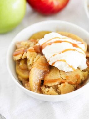 Easy apple cobbler topped with whipped cream and caramel sauce in a white bowl.