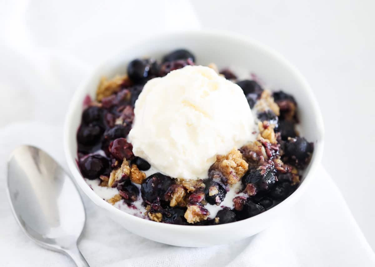 An easy blueberry crisp with a thick sweet filling topped with a toasty cinnamon sugar streusel topping is the best way to welcome cooler weather. It's a quick dessert or afternoon snack that everyone finds comfort in! Blueberry crisp with ice cream in a white bowl.