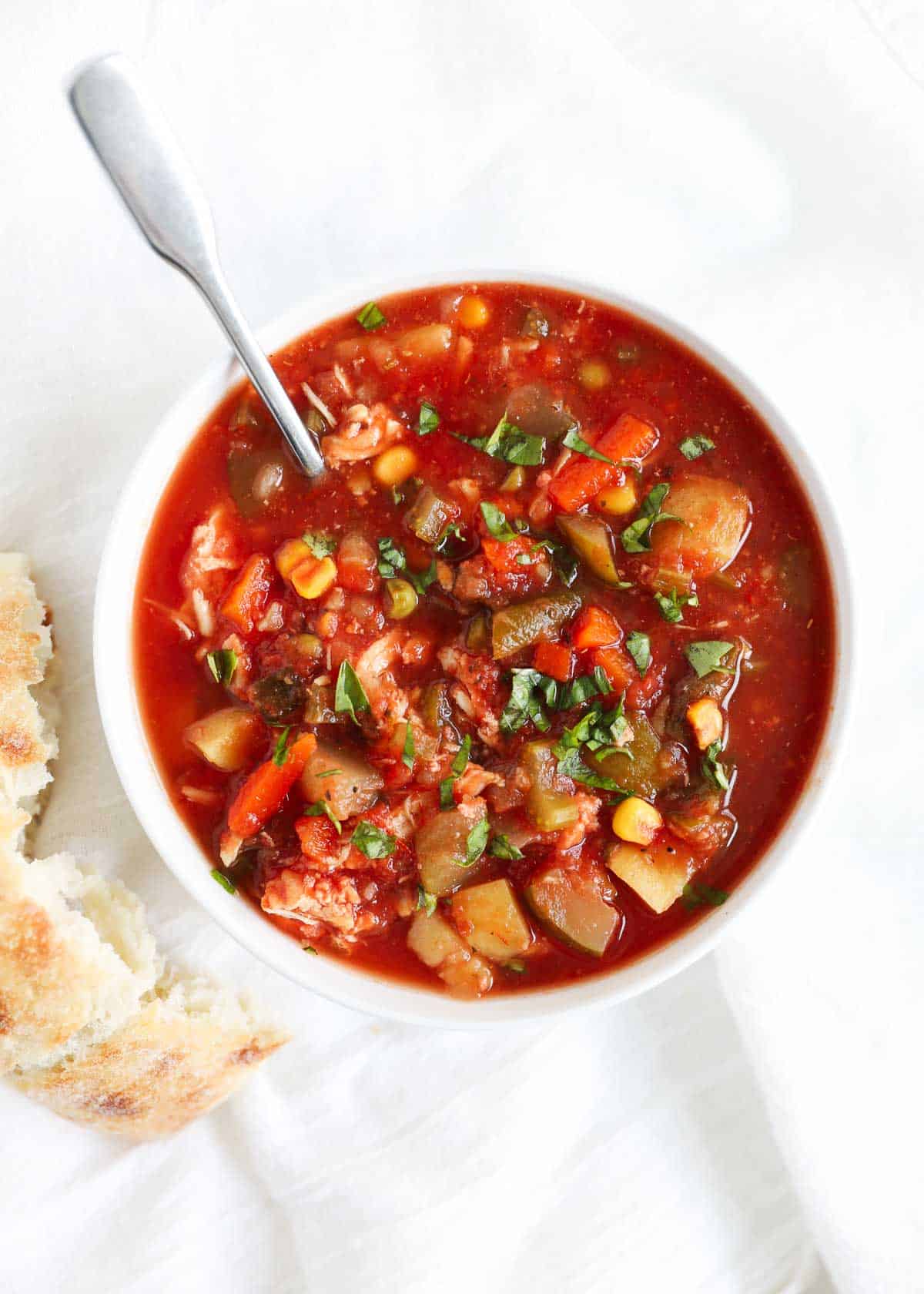 Chicken vegetable soup in a bowl with spoon and crusty bread on the side.