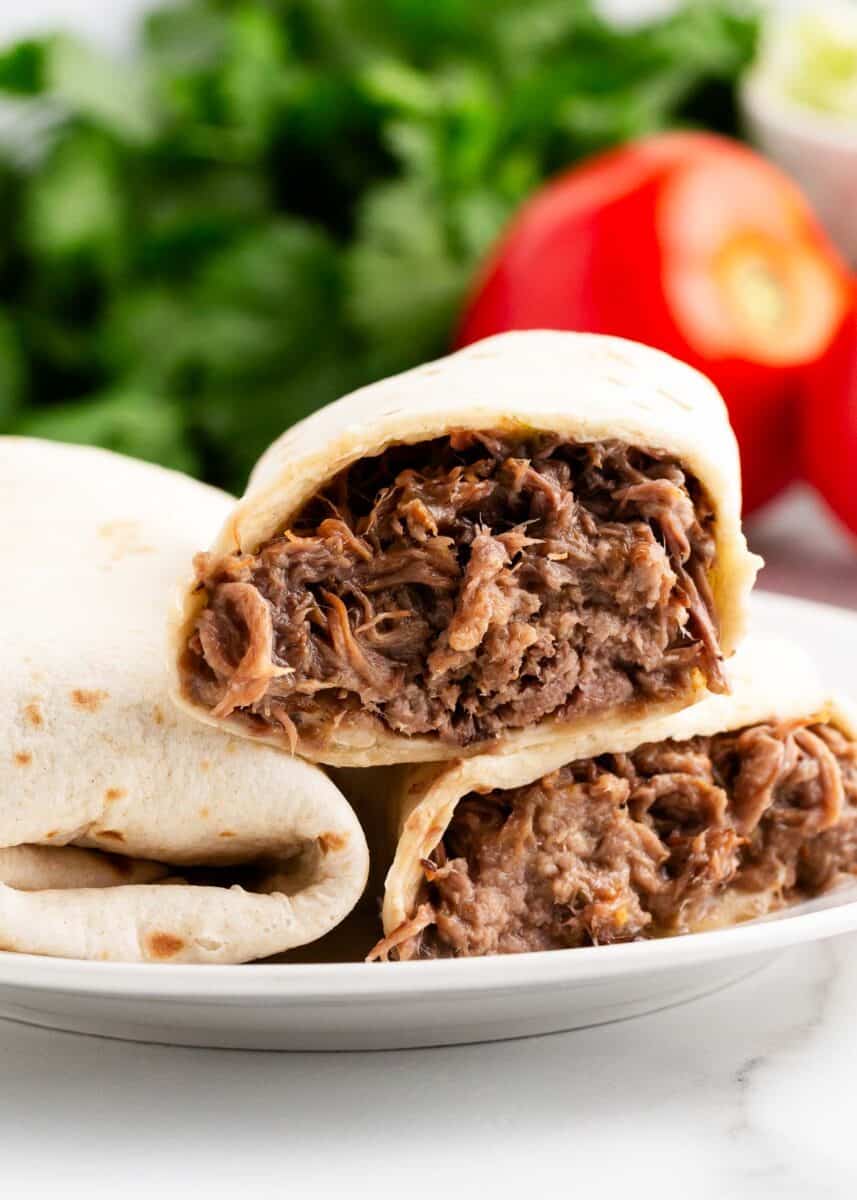 Warm flour tortillas filled with tender slow cooker shredded beef and finished off with your favorite toppings. These green chili burritos are delicious, easy, and perfect for a weeknight dinner for busy families! stacked green chili burritos on white plate