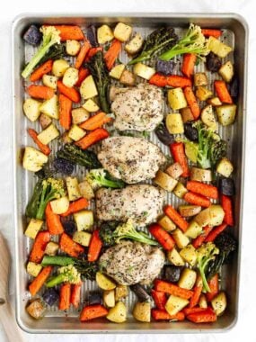Overhead view of baked sheet pan chicken thighs with roasted carrots, potatoes, broccoli, and purple potatoes, colorful and arranged neatly for serving.