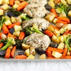 Angled view of baked sheet pan chicken thighs surrounded by roasted carrots, potatoes, broccoli, and purple potatoes, the vegetables are tender with lightly browned edges.