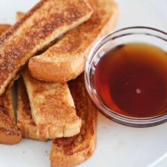 Homemade cinnamon French toast sticks on a plate with a bowl of maple syrup for dipping.