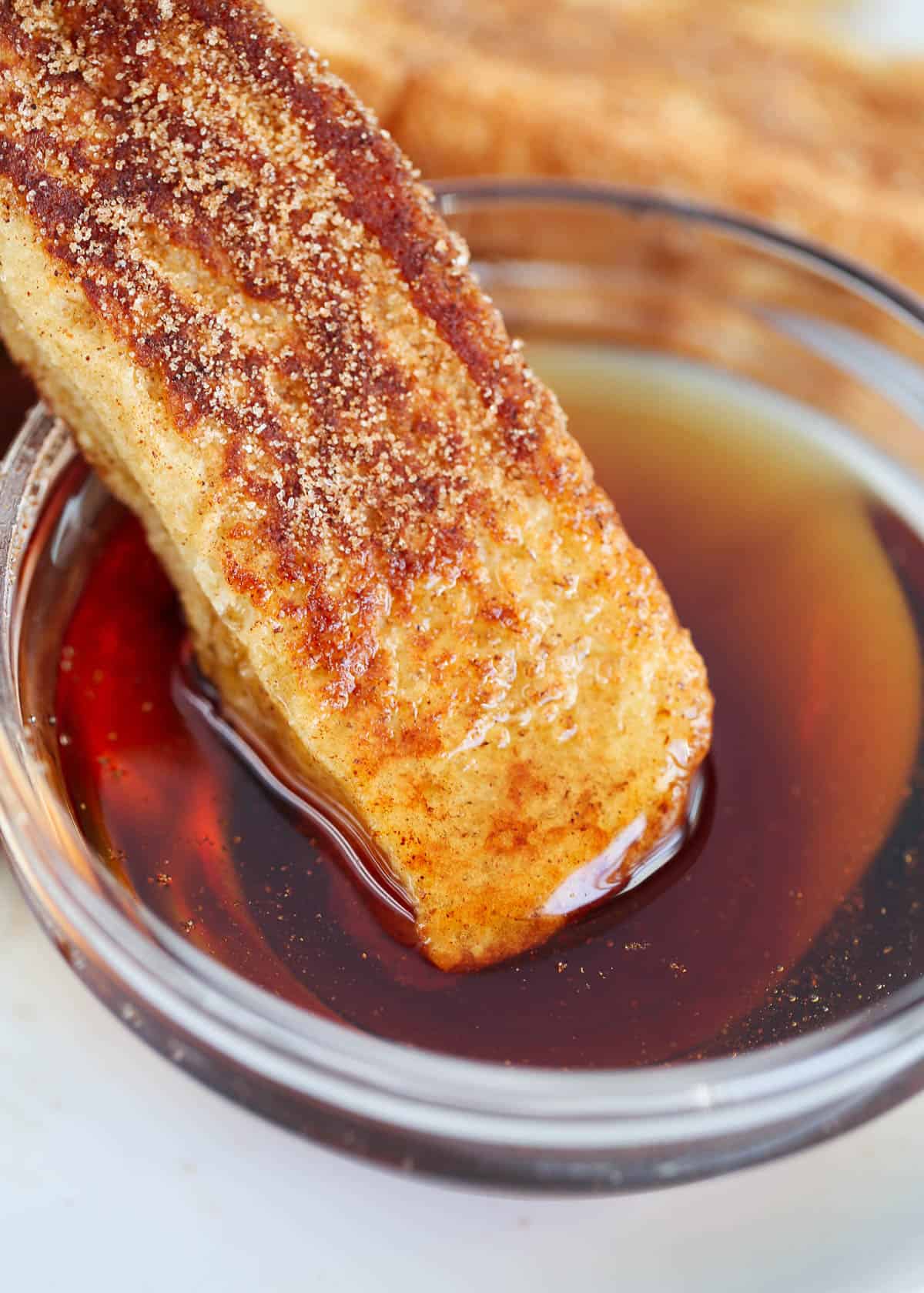 French toast sticks are fun for all ages! Strips of thick pieces of toast are made into cinnamon-flavored dippers perfect for dipping in your favorite syrup, whipped cream, or sprinkled in a little extra sugar! Dipping cinnamon French toast stick in syrup.
