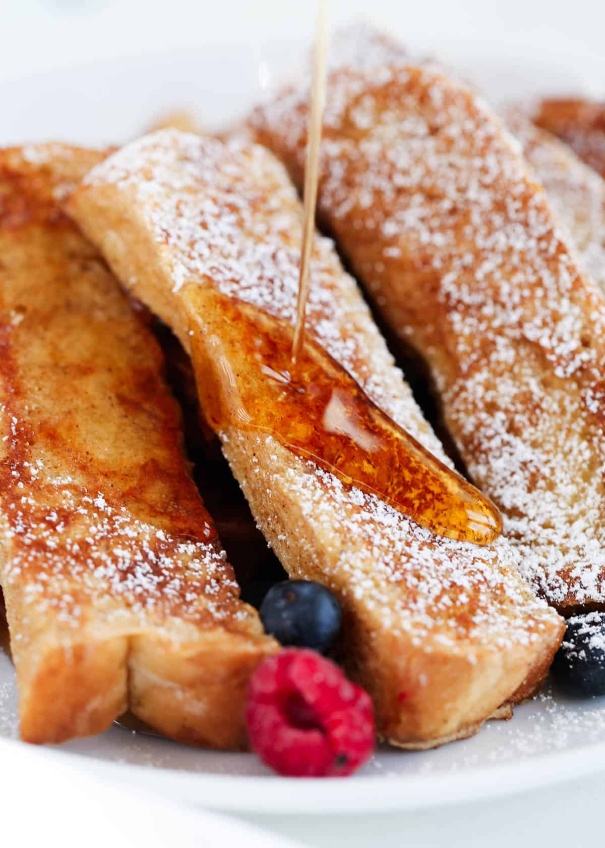 French toast sticks are fun for all ages! Strips of thick pieces of toast are made into cinnamon-flavored dippers perfect for dipping in your favorite syrup, whipped cream, or sprinkled in a little extra sugar! Pouring syrup over homemade French toast sticks dusted with powdered sugar and served with berries.