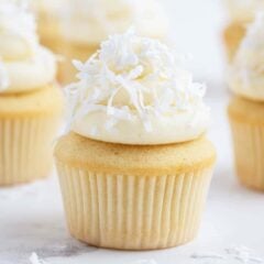 Coconut cupcakes on the counter topped with fluffy coconut frosting and coconut flaikes.