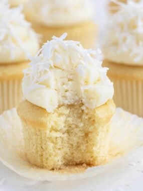 Coconut cupcake with a bite taken out, topped with coconut cream cheese frosting and shredded coconut.