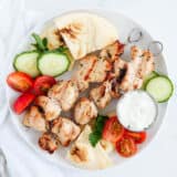 Chicken kabobs on a plate with pita bread, sliced tomatoes and cucumbers.