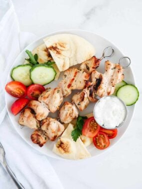 Chicken kabobs on a plate with pita bread, sliced tomatoes and cucumbers.
