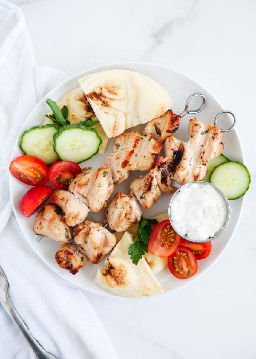 Chicken kabobs on a plate with pita bread, sliced tomatoes and cucumbers.