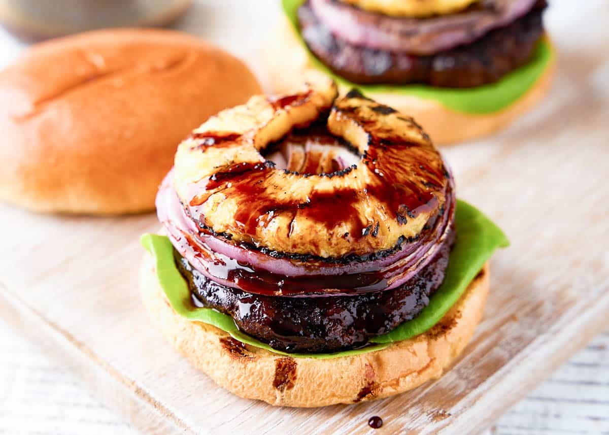 This teriyaki burger recipe is a must-try! Juicy burger flavored with a sweet teriyaki sauce and served with grilled pineapple and onions on a crispy brioche bun! Open teriyaki burger with onion and pineapple on top on wooden board.