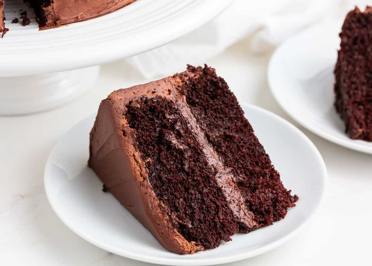 It's a rich, moist chocolate cake layered with chocolate buttercream frosting all made from scratch using one bowl and pure and simple ingredients! Slice of chocolate cake on white plate.
