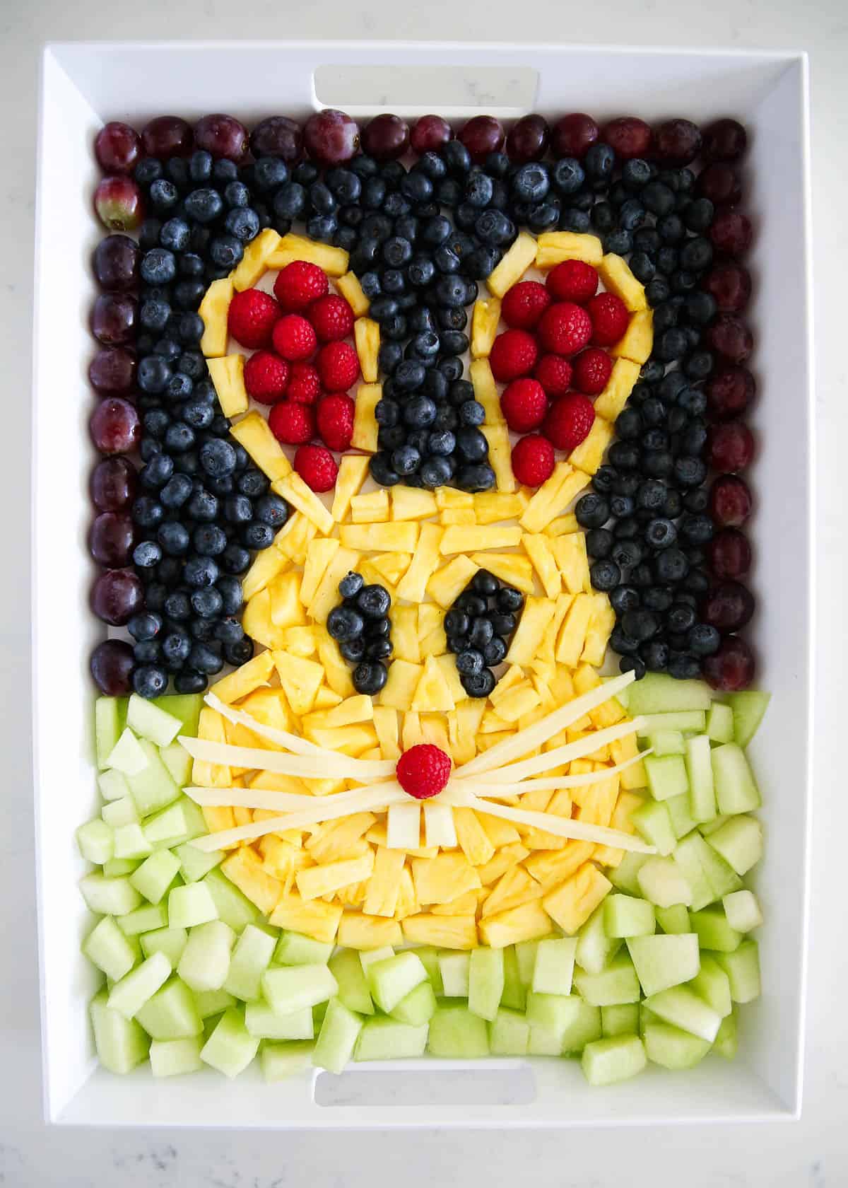A white platter with berries, pineapple and melon in the shape of a bunny rabbit. 