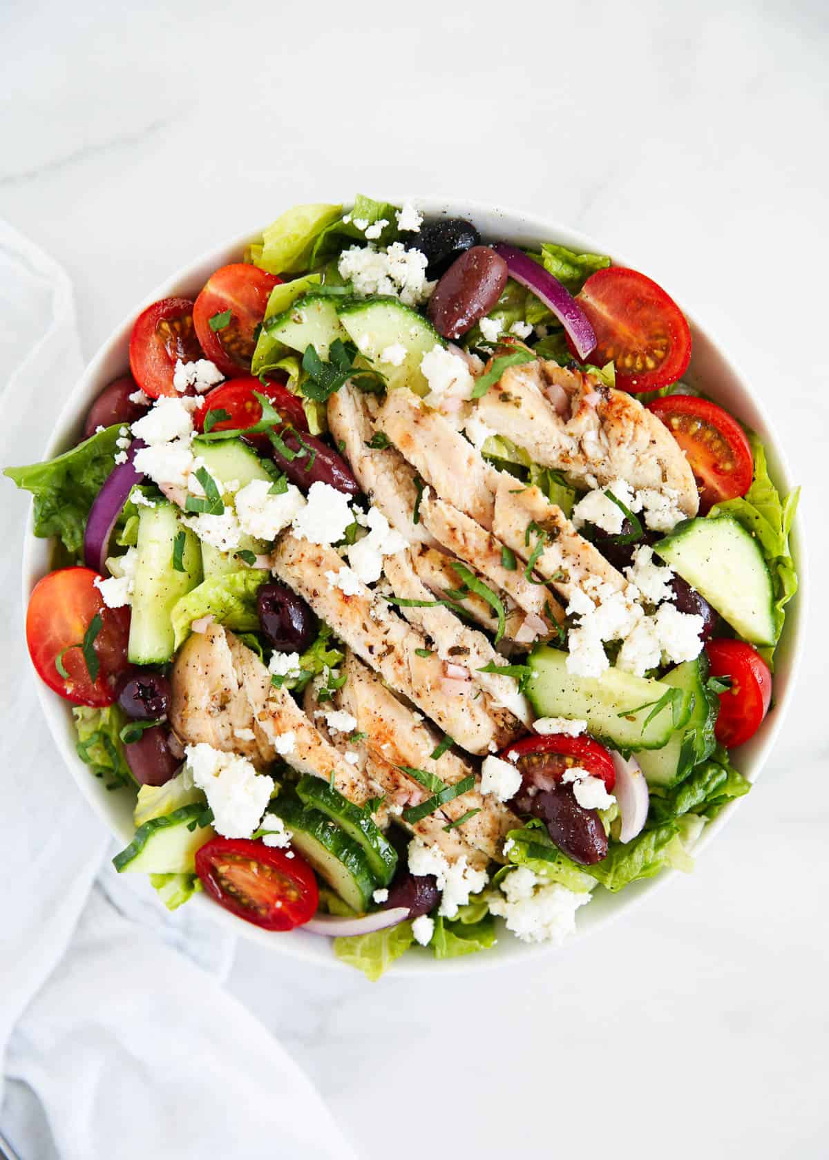 This Greek salad with chicken is a protein-packed twist on a classic Greek salad! It's loaded with fresh crispy veggies, juicy tomatoes, briny feta, and kalamata olives and topped with slices of grilled chicken! Greek chicken salad in a white bowl on the counter with a white linen.