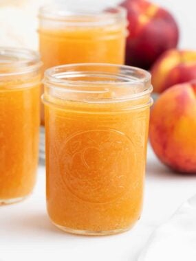 Three jars of peach freezer jam on the counter with fresh peaches nearby.