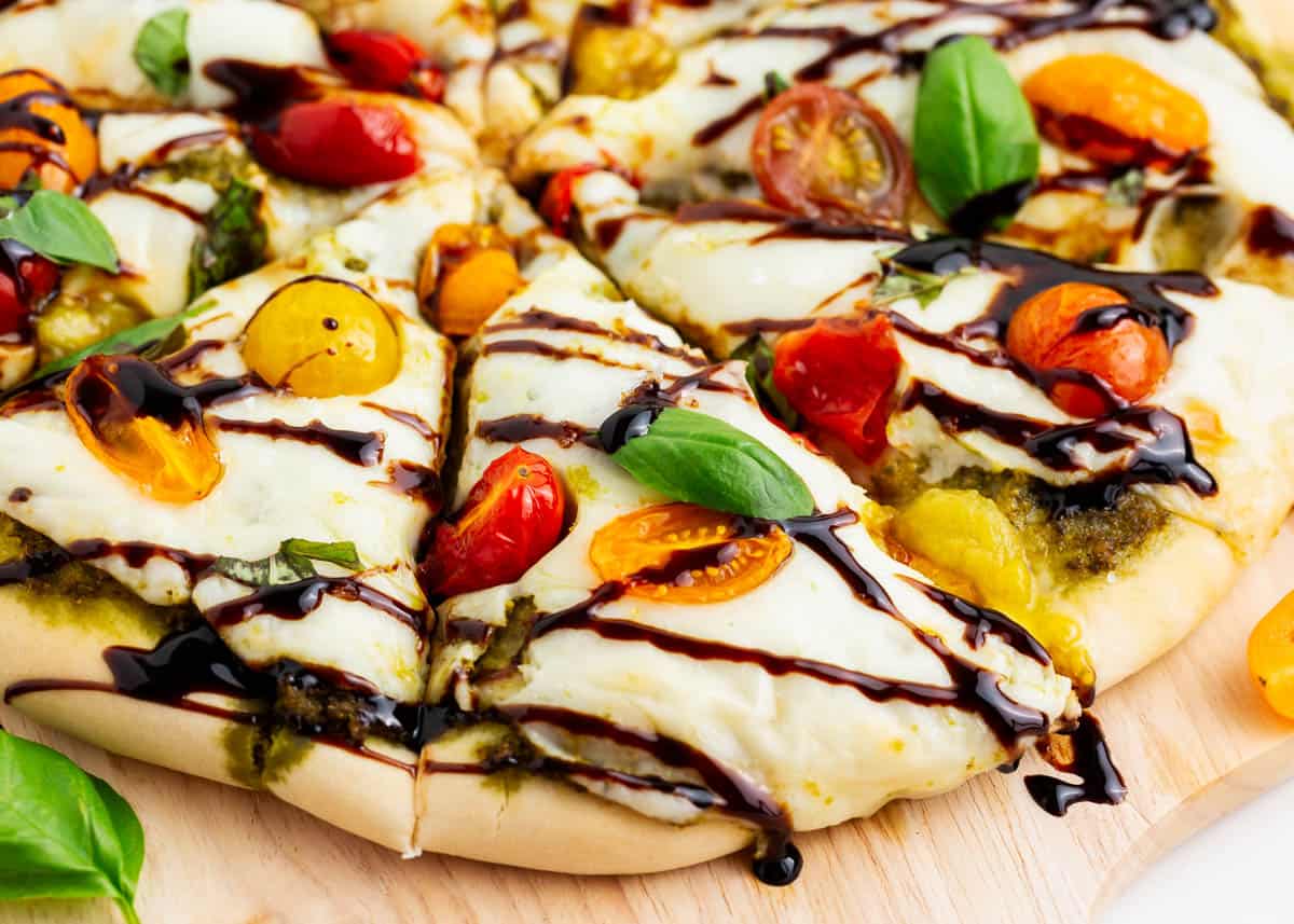 Pizza Caprese is made with a crispy golden crust topped with basil pesto, gooey mozzarella, zesty tomatoes, and fresh basil leaves. One bite is enough to transport you to an Italian pizzeria! Sliced caprese pizza drizzled with a balsamic glaze.