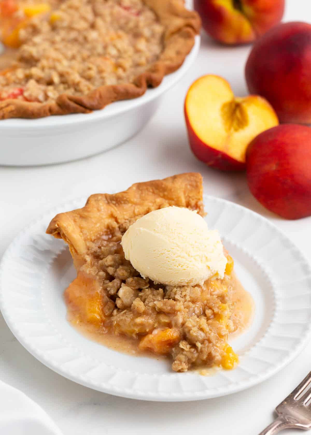 Slice of peach pie with crumb topping and a scoop of vanilla ice cream.