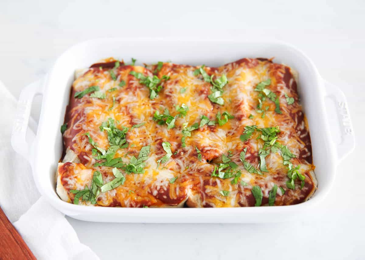 Pork enchiladas are a delicious, filling, and flavorful Mexican-inspired dish! They're filled with seasoned pulled pork, beans, cheese, and a smoky enchilada sauce. Baked to perfection, this dish has been a longtime family favorite for a reason! Pork enchiladas in white baking dish on counter.