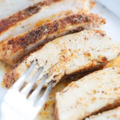 A piece of juicy baked chicken breast sliced on a white plate with a fork.
