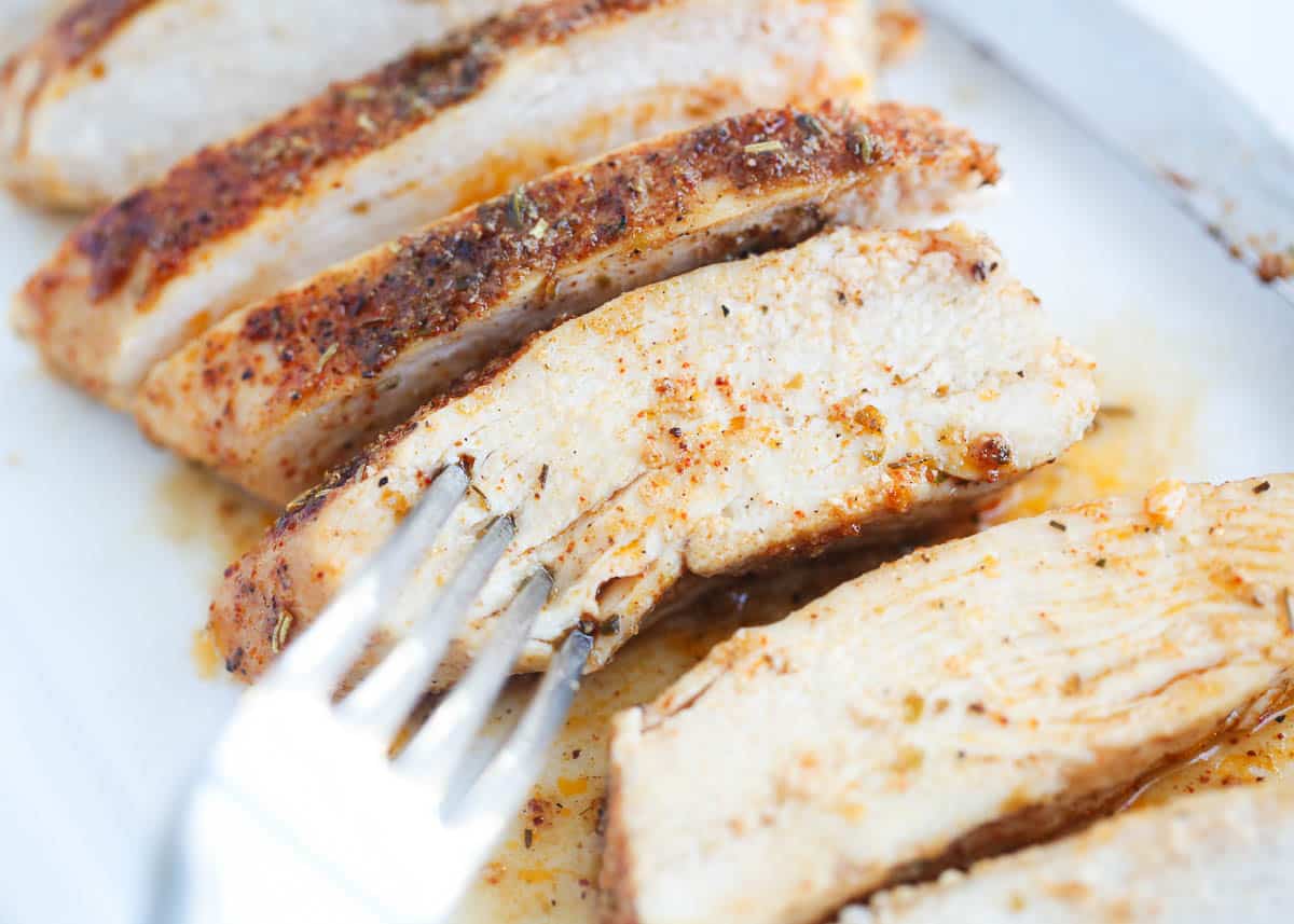 This is the juiciest and most flavorful easy oven baked chicken breast recipe! It's made with or without a brine, a handful of simple seasonings, then baked to perfection every time! Sliced baked chicken breast on a white plate with a fork.