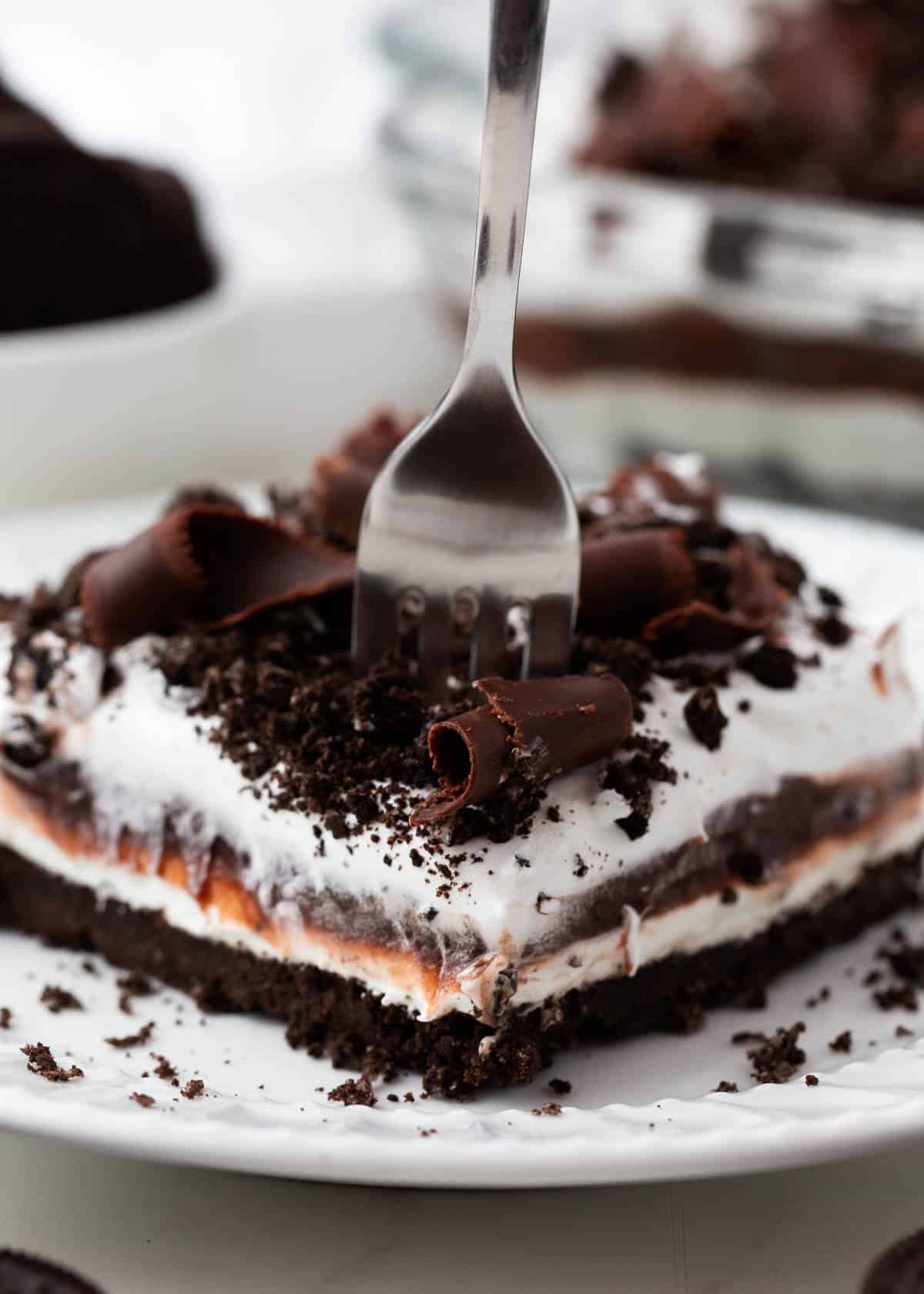 Fork taking a bite out of a chocolate lasagna slice, show layers of Oreo crust, creamy pudding and whipped topping.