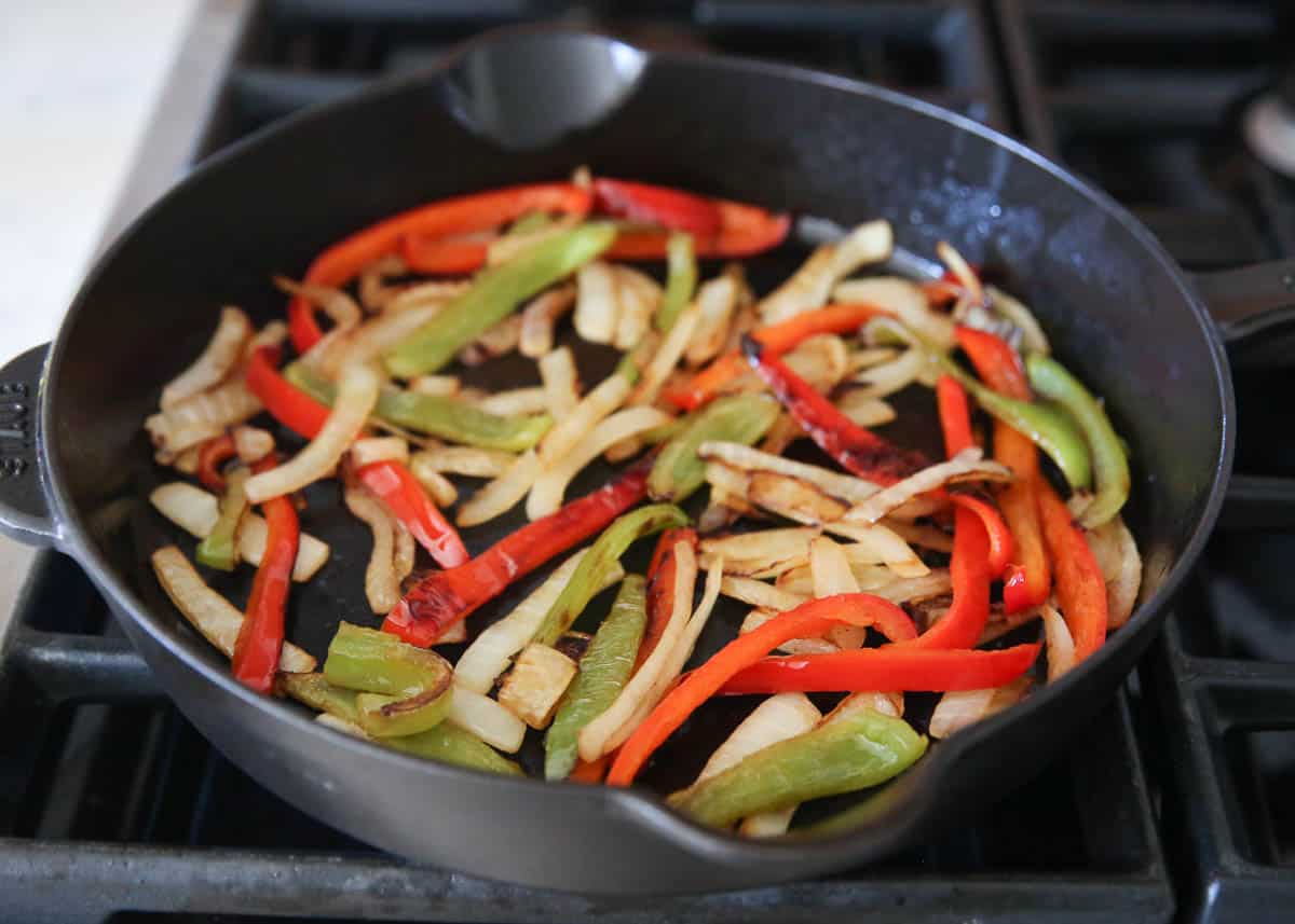 Caramelized red and green bell peppers and onions cooked down in a black cast iron skillet on a gas stovetop.