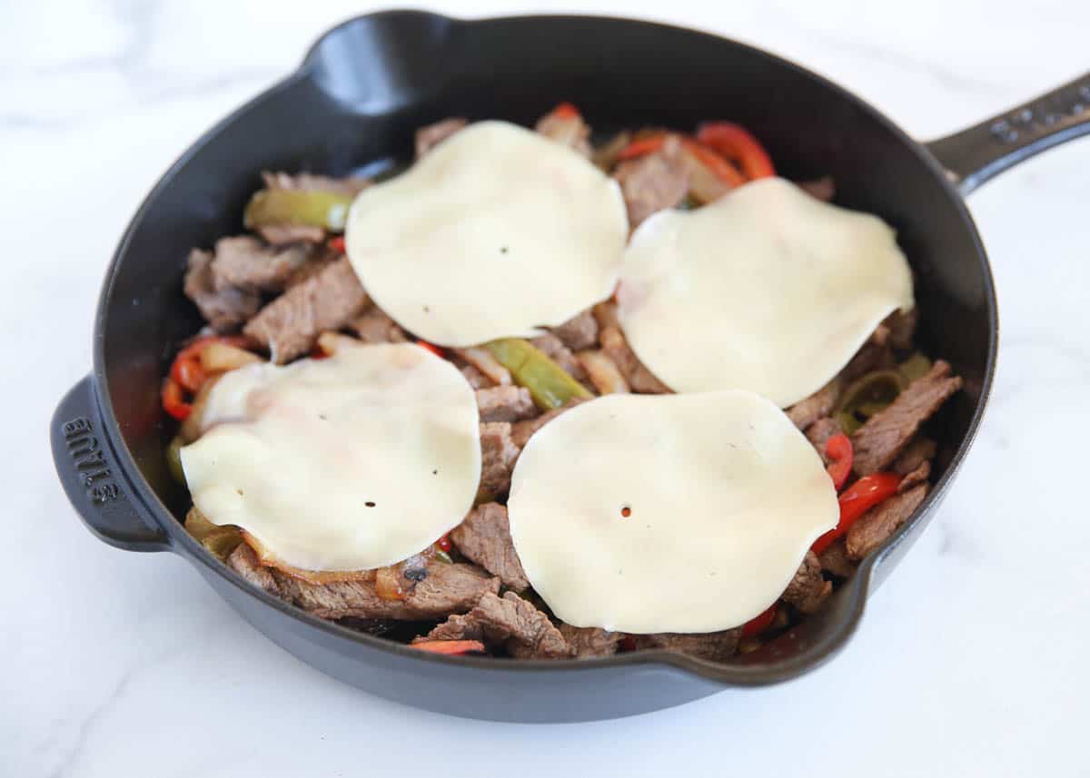 Four slices of melted provolone cheese draped over cooked ribeye steak, peppers and onions in a black Staub cast iron skillet.