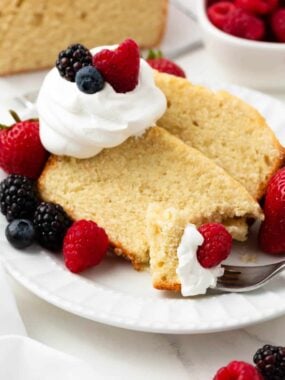 Two slices of classic pound cake on a white plate surrounded by fresh strawberries, raspberries, blueberries, and blackberries with a dollop of whipped cream.