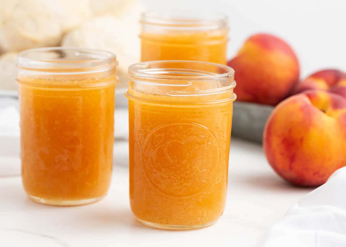 Three jars of freshly made peach freezer jam with ripe peaches in the background.