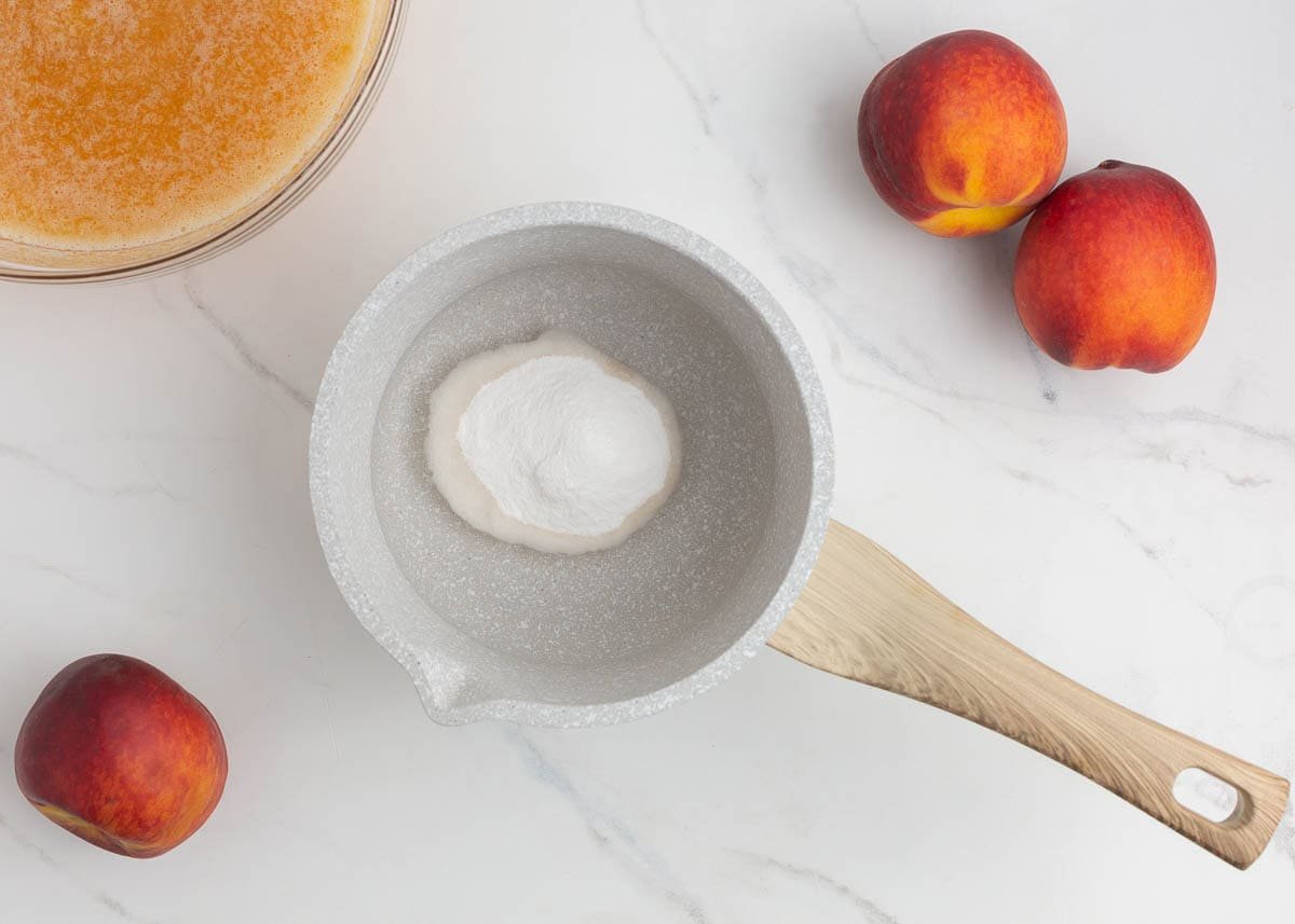 Powdered pectin in a saucepan, surrounded by peaches and prepared peach puree.