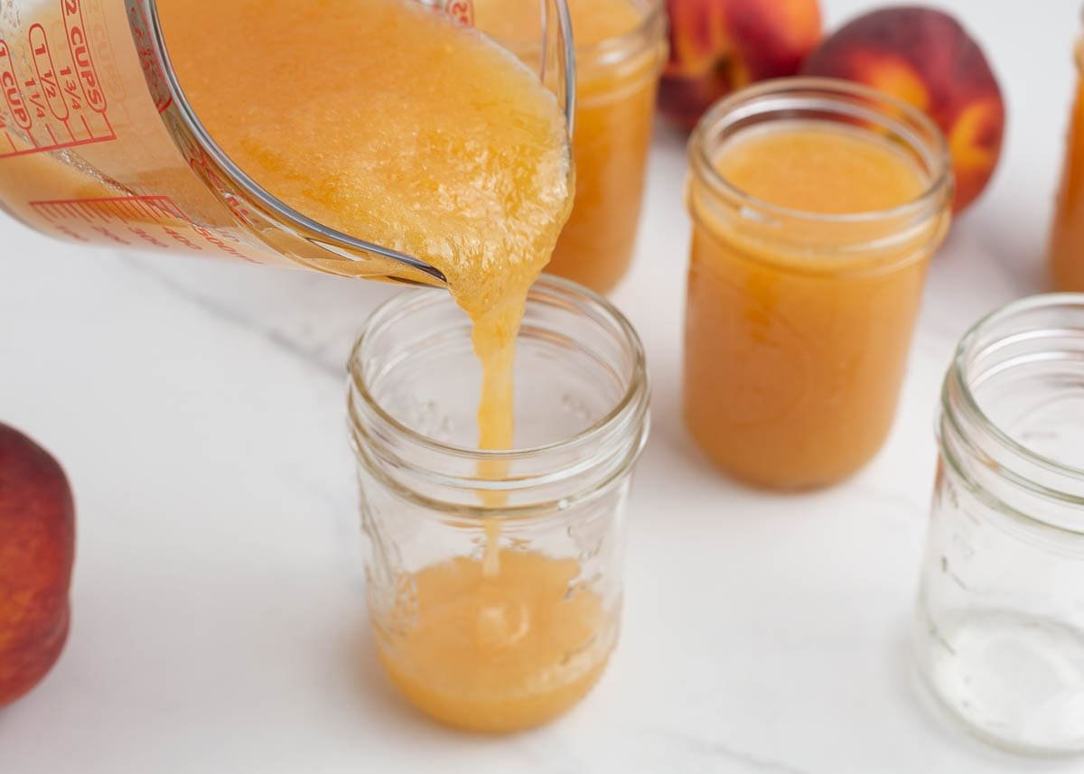 ouring homemade peach freezer jam into a jar using a measuring cup.