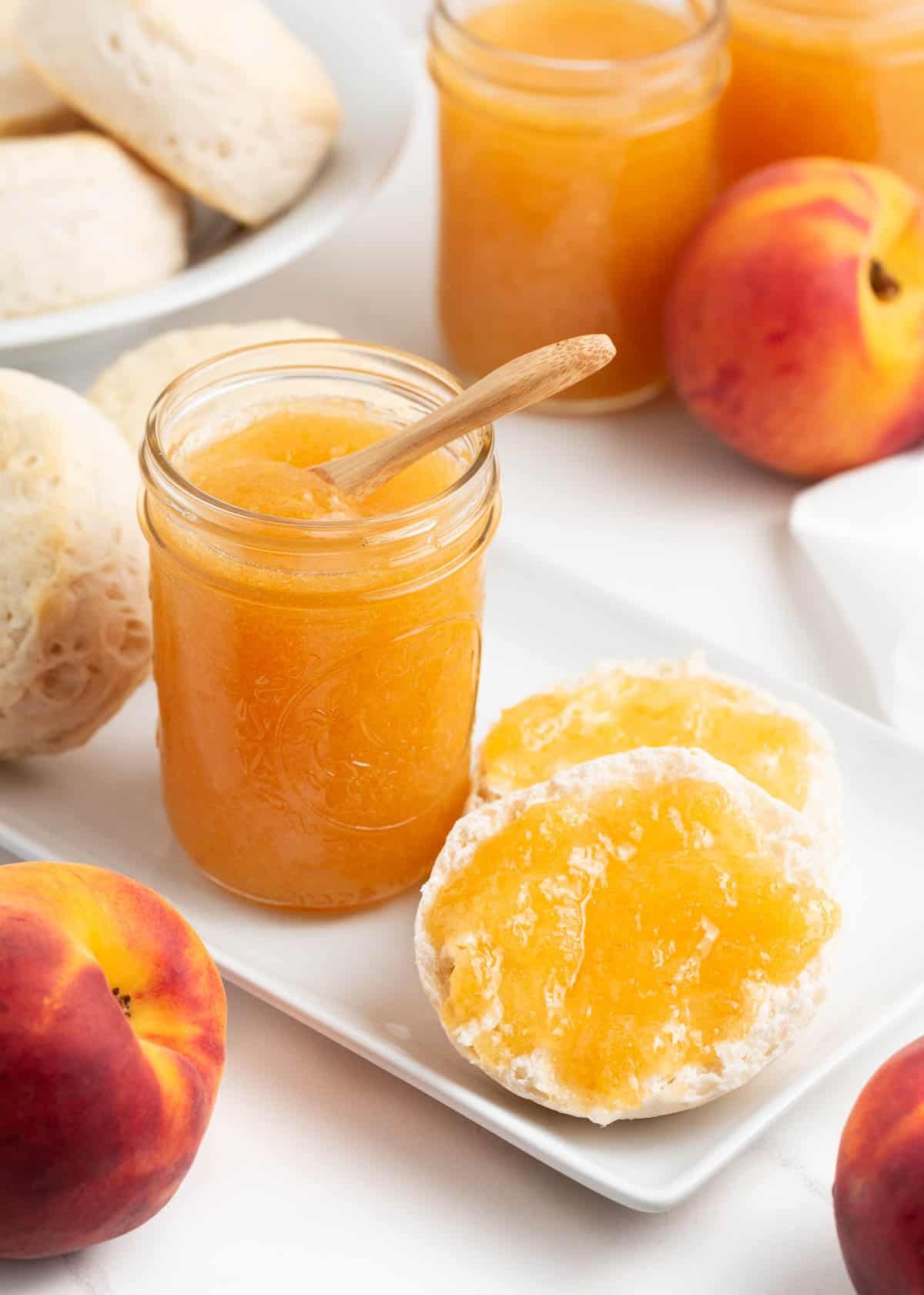 Open jar of peach freezer jam with a spoon, served next to biscuits and fresh peaches.