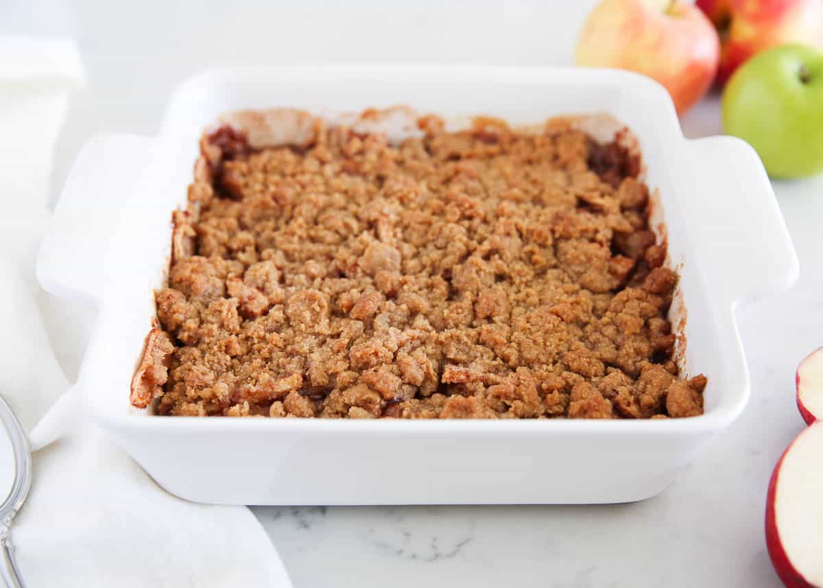 This apple crumble recipe is easy to make with a tender apple filling baked under a buttery brown sugar crumble topping. It's our favorite apple dessert for fall! Apple crumble recipe baked in a white baking dish with fresh apples on counter.