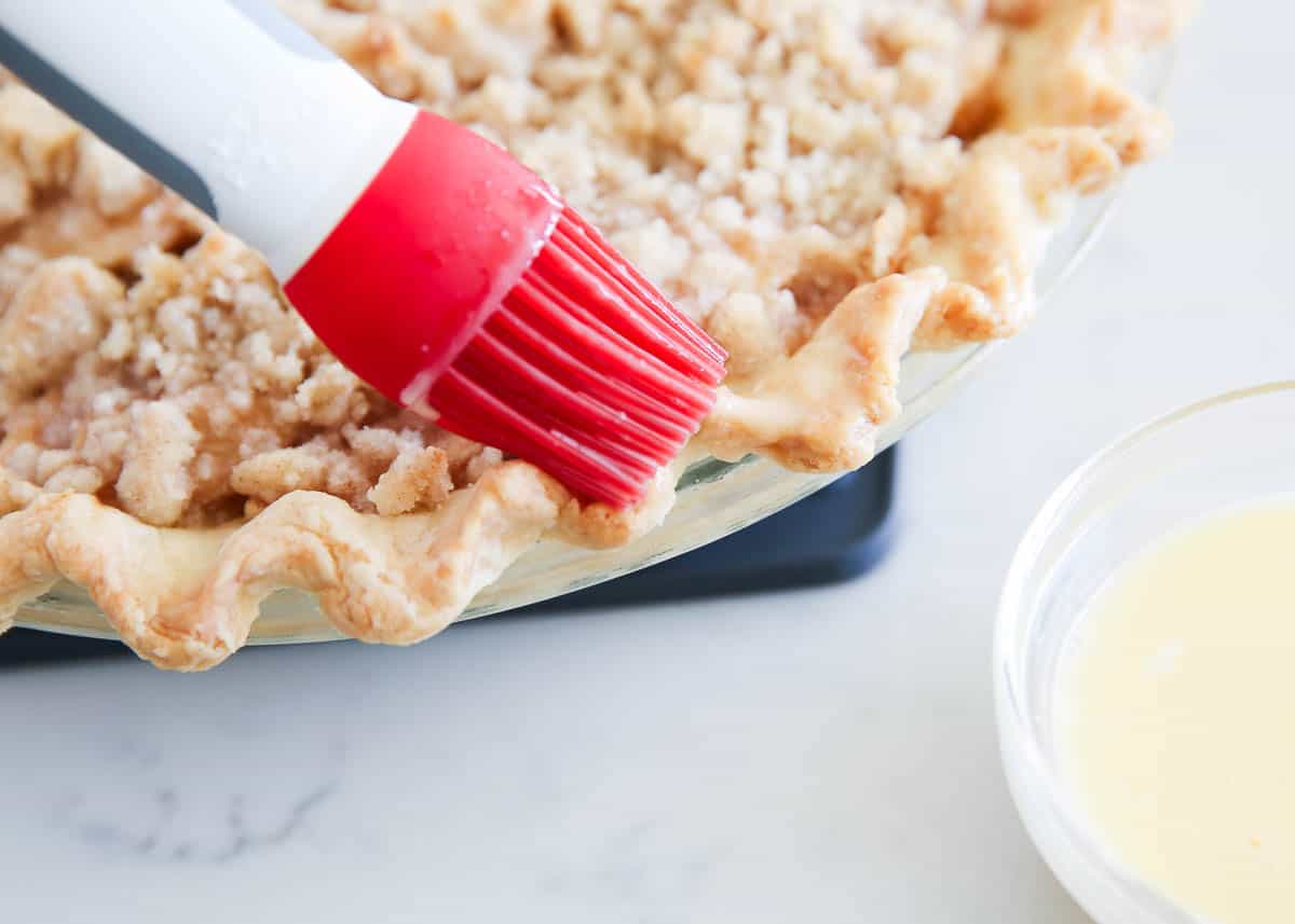 Learn how to use this easy egg wash recipe to create beautiful finishes on your bakes! This technique works for pastry dough, and so much more! The results are an irresistible golden brown color and shiny crust. Brushing egg wash onto pie crust.
