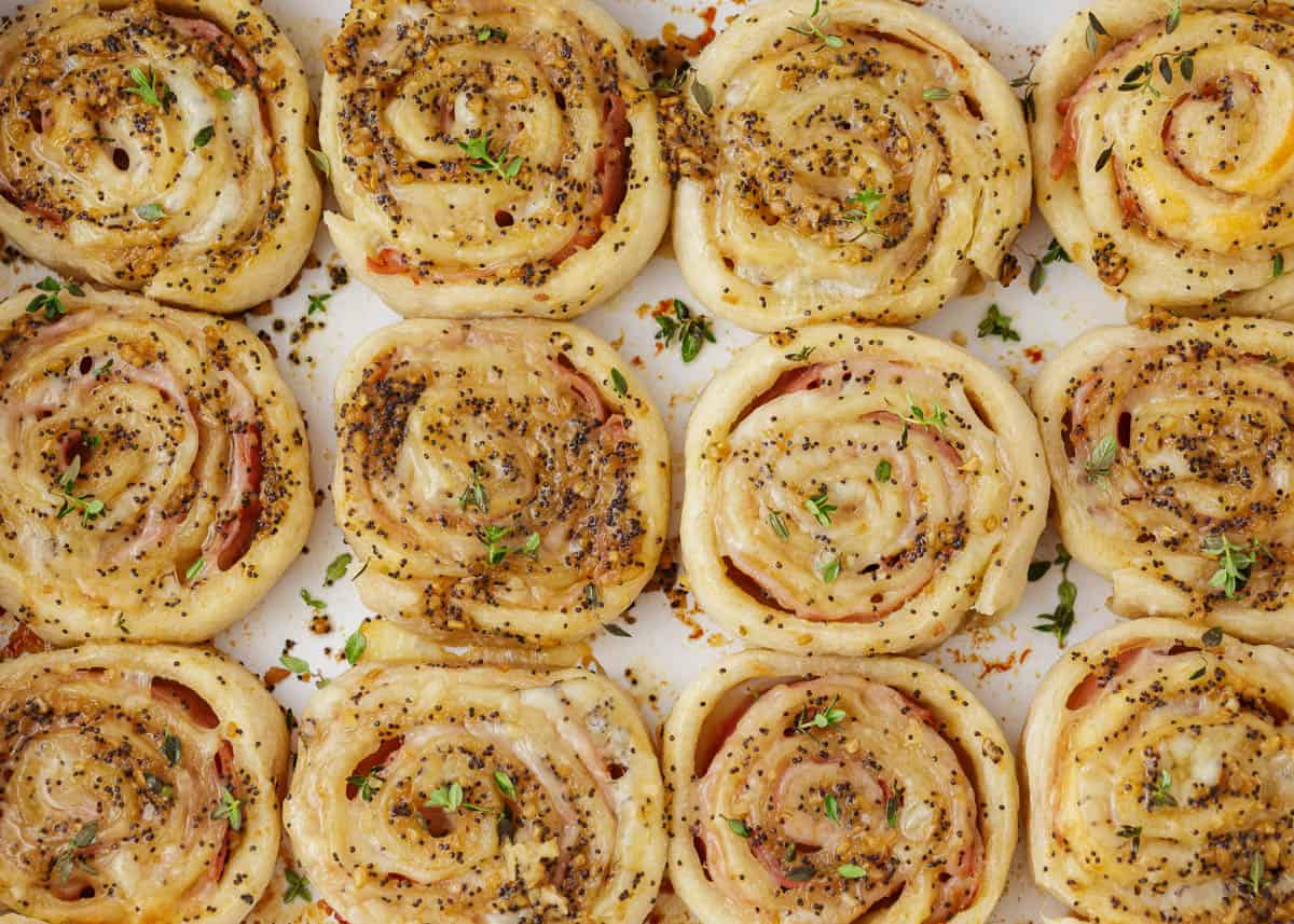 Crispy rolls filled with melty cheese, and savory ham, brushed with a Dijon glaze and topped with poppy seeds are what make these baked ham and cheese pinwheels so tasty! A popular appetizer or snack made in under 30 minutes! Ham and cheese pinwheels in a baking dish.