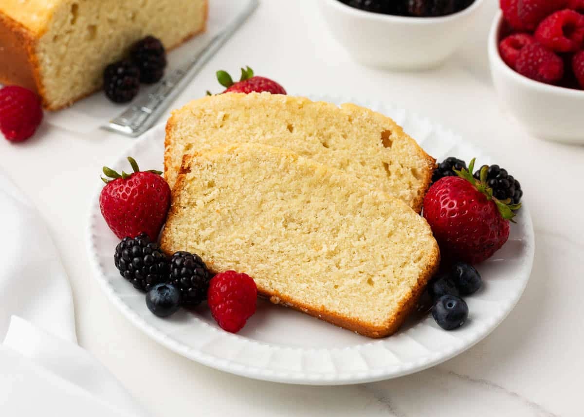 Two slices of classic pound cake on a white plate surrounded by fresh strawberries, raspberries, blueberries, and blackberries.