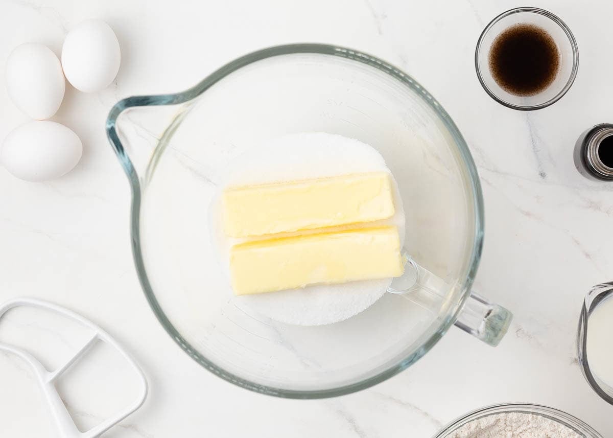 Butter and sugar in a stand mixer bowl with eggs, vanilla, milk, and flour nearby.