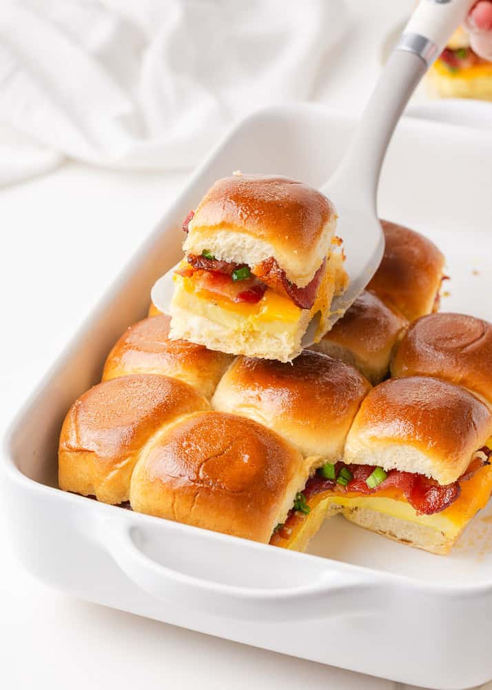 Wow your breakfast or brunch crowd with these crispy golden buns filled with bacon, eggs, and cheddar cheese! Baked as one, then pulled apart, these Hawaiian roll breakfast sliders are easy to make and even easier to serve! A spatula lifting one Hawaiian roll breakfast slider out of a white baking dish filled with a dozen golden-baked breakfast sliders.