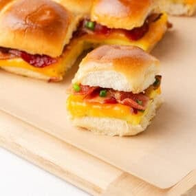 Close-up of a single Hawaiian roll breakfast slider on a wooden cutting board, showing layers of fluffy scrambled egg, melted cheddar cheese, crispy bacon, and green onions, with more sliders in the background.