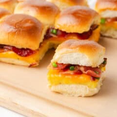 A pulled-apart Hawaiian roll breakfast slider on a wooden board showing layers of scrambled eggs, melted cheddar, crispy bacon, and sliced green onions, with more assembled sliders behind it.