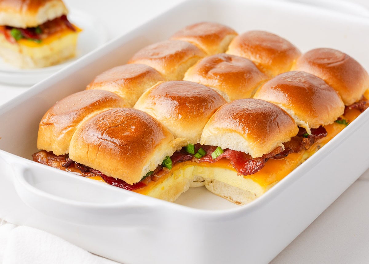 Hawaiian roll breakfast sliders in a white baking dish with melted cheddar cheese, crispy bacon, and green onions visible between the golden glazed buns.